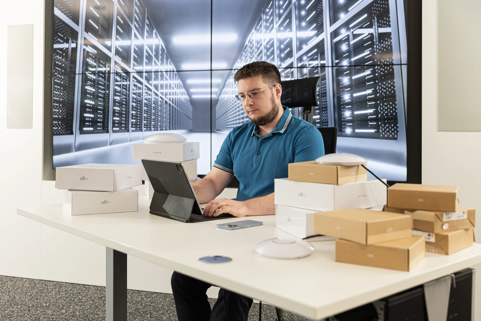 Mann an Schreibtisch arbeitet am Laptop, umgeben von Routerboxen, mit großem Bildschirm im Hintergrund, der Serverraum zeigt.
