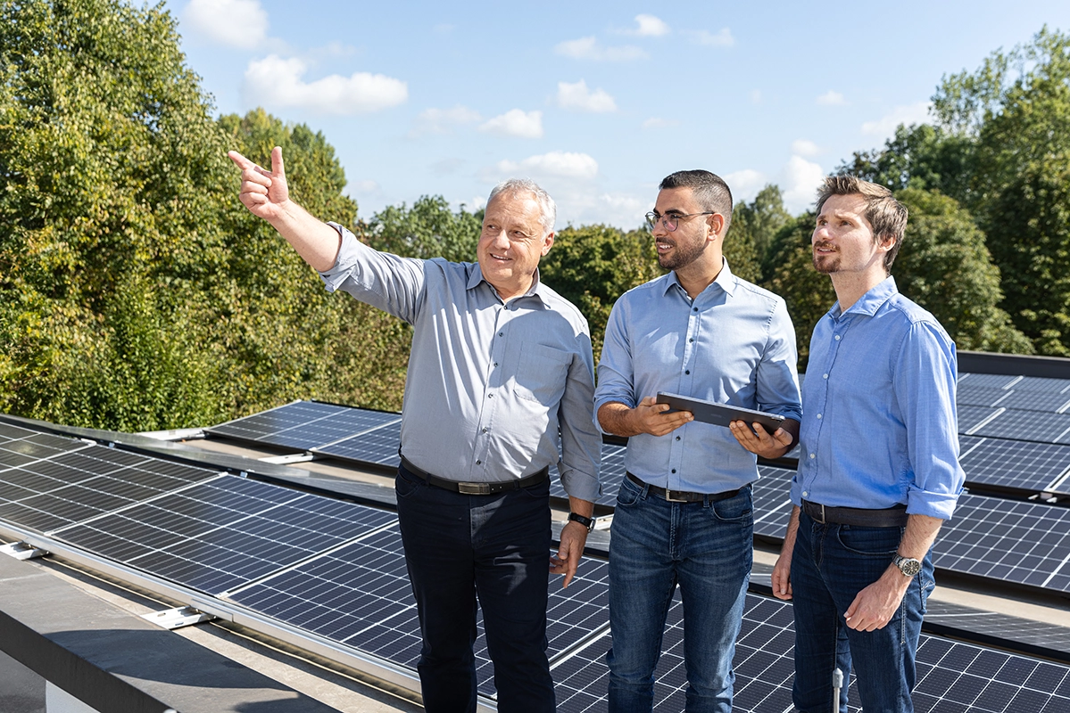 Drei Männer stehen auf einem Dach voller Solarpanels, einer zeigt in die Ferne, ein anderer hält ein Tablet.
