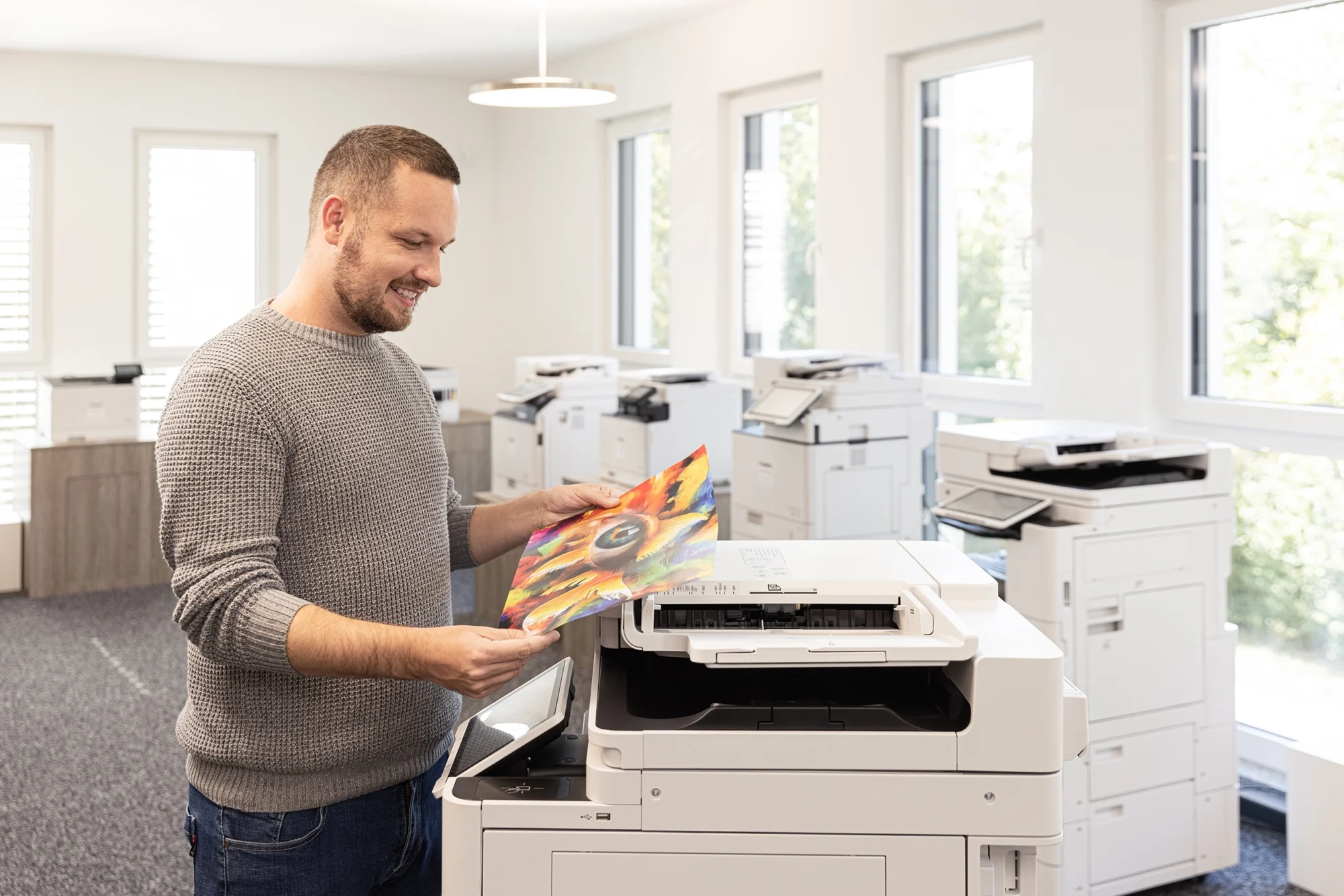 Ein Mann in grau, hält ein buntes Druckbild in einem hellen Büro, umgeben von mehreren Druckern.