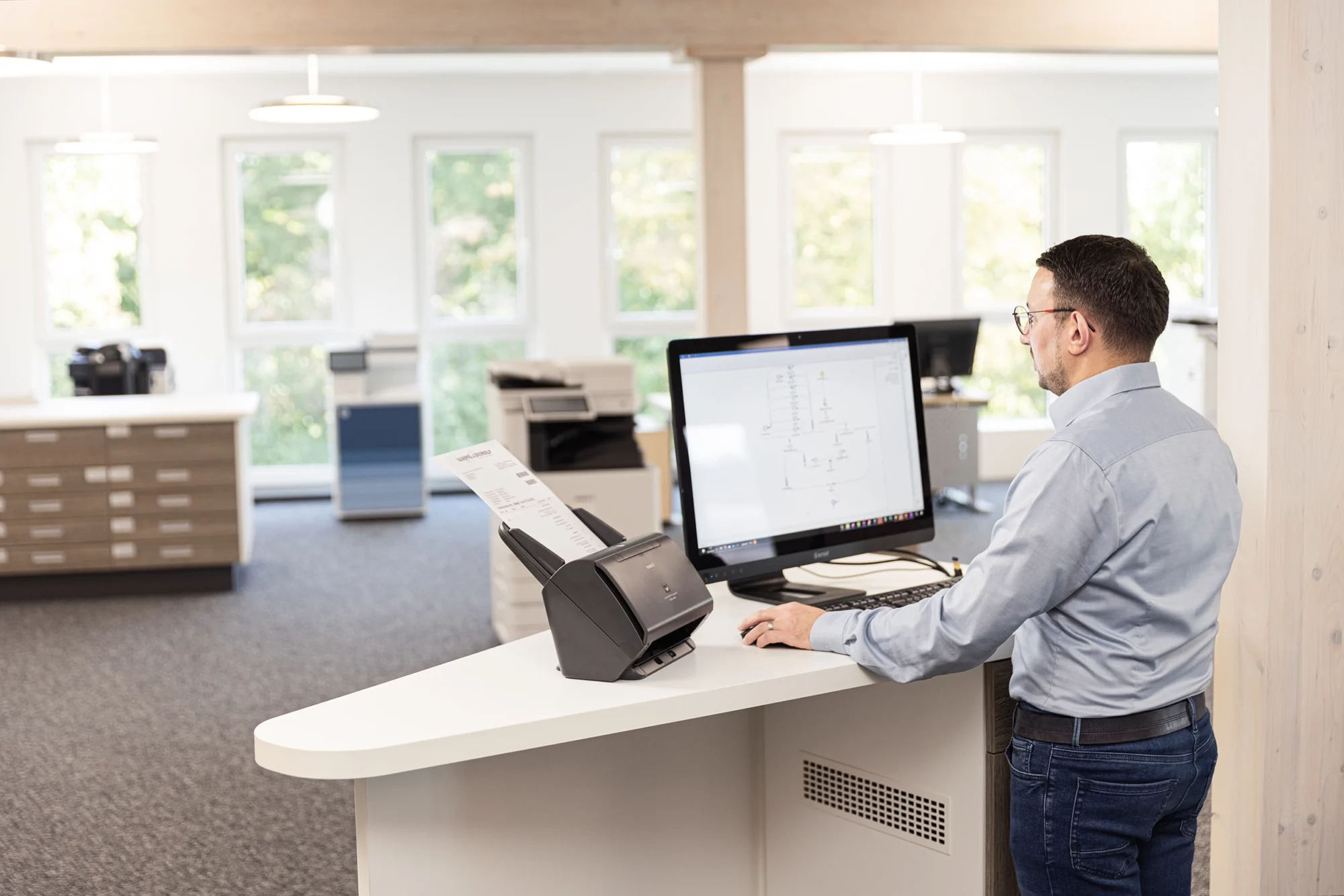 Mann im Büro arbeitet an Computer mit Scanner am Schreibtisch, helle Fenster im Hintergrund.