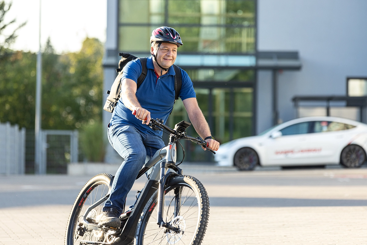 Person mit Helm fährt lächelnd auf einem E-Bike vor einem modernen Gebäude mit Glasfassade. Hintergrund zeigt ein weißes Auto.