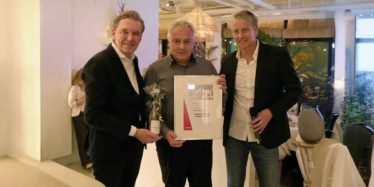 Drei Männer in Anzügen halten eine Urkunde und eine Flasche in einem elegant beleuchteten Raum in die Kamera.