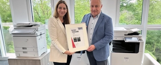 Zwei Personen halten eine Urkunde in einem Büro mit zwei Druckern im Hintergrund.