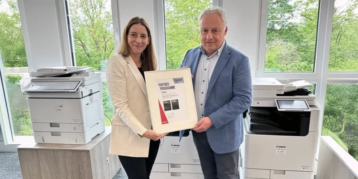 Zwei Personen halten eine Urkunde in einem Büro mit zwei Druckern im Hintergrund.