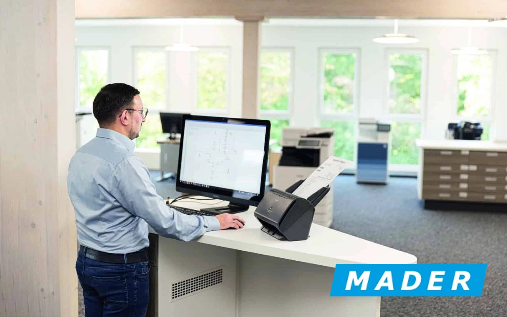Kappel-Dierolf_Cases_Mader Mitarbeiter steht vor Computer in modernem Büro, nutzt Scanner für Dokumenteneingabe. Firmenlogo im Vordergrund.