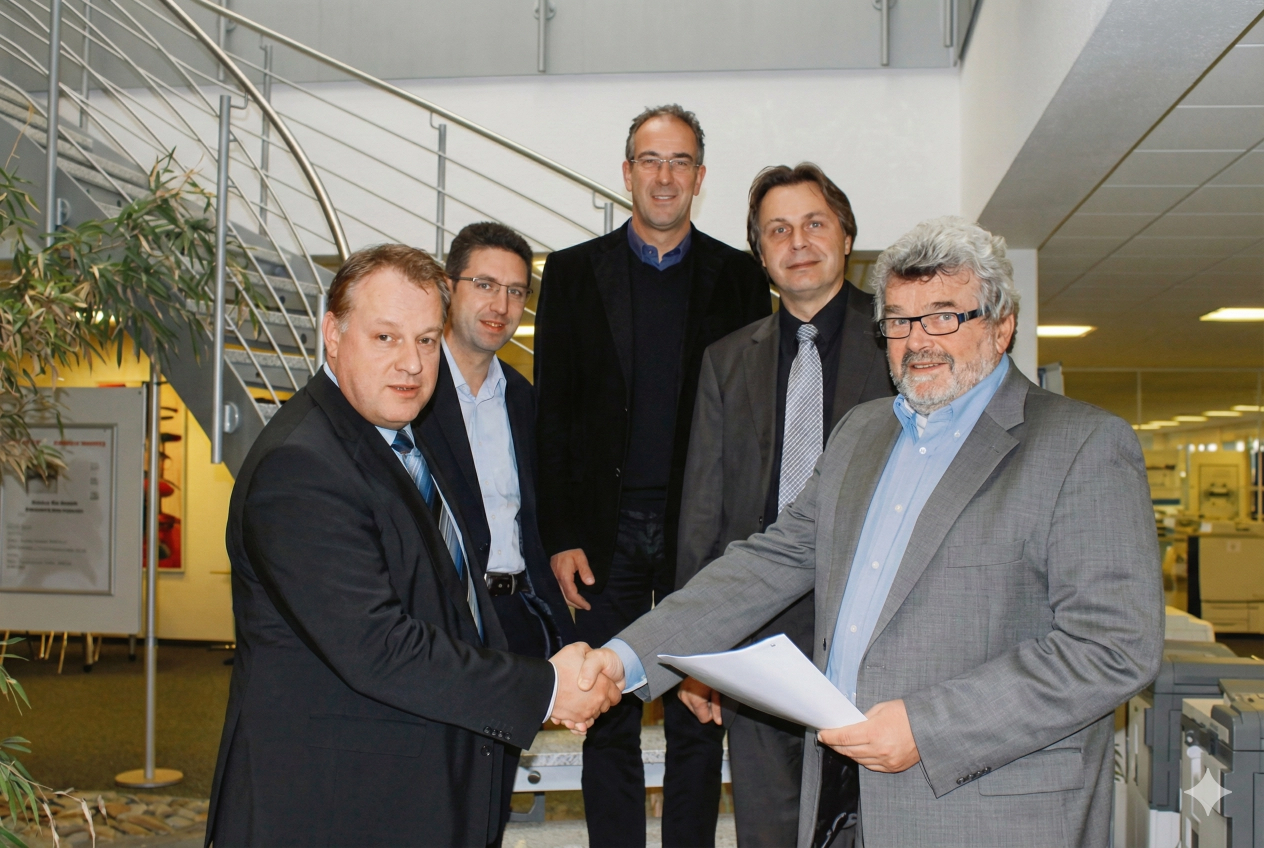 Fünf Männer in Anzügen stehen zusammen. Zwei Männer im Vordergrund schütteln die Hände und halten ein Dokument.