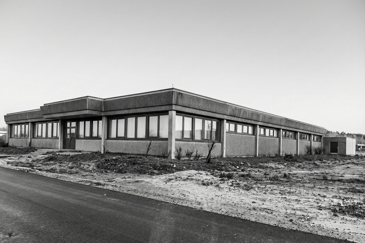 Graues, rechteckiges Betongebäude mit vielen Fenstern und einer Eingangstür, umgeben von karger Landschaft.