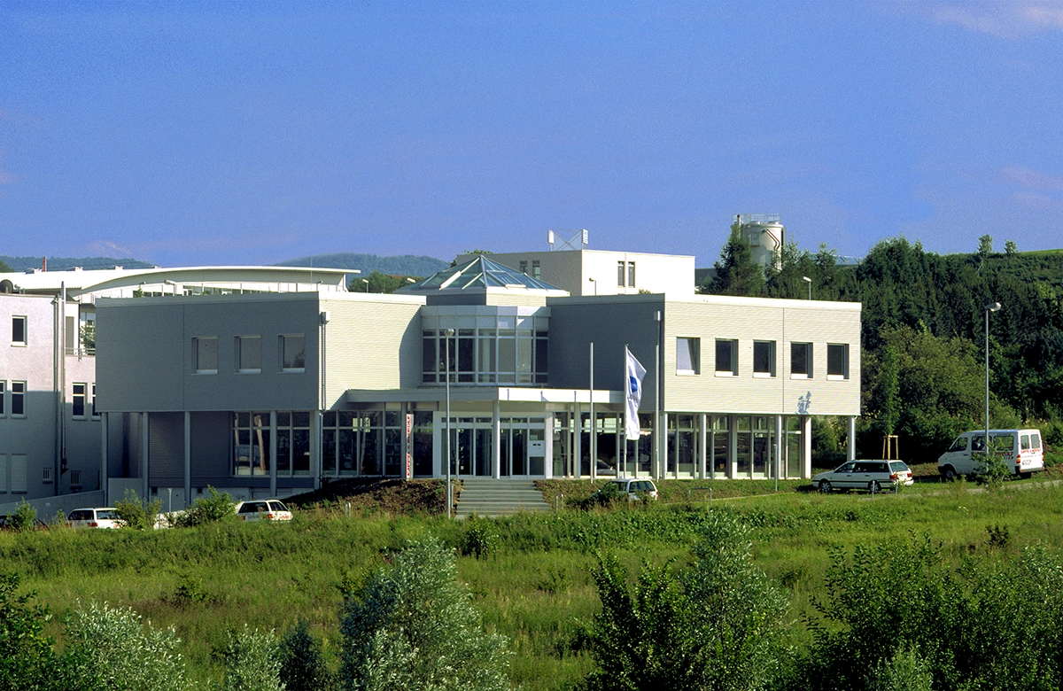 Modernes Bürogebäude mit Glasdach und mehreren Fahrzeugen davor, umgeben von grüner Vegetation und klarem Himmel.