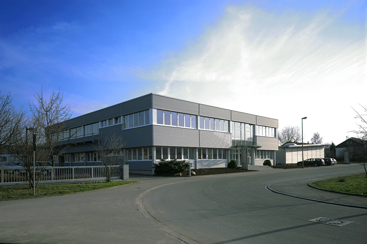 Modernes zweistöckiges Bürogebäude mit großen Fenstern, umgeben von einer Straße und kahlen Bäumen unter klarem Himmel.