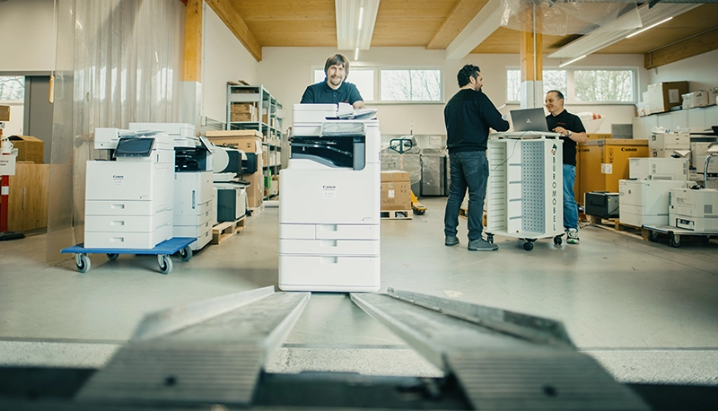 Drei Männer arbeiten in einem Büro mit Druckern und Kopierern, umgeben von Verpackungen und Regalen.