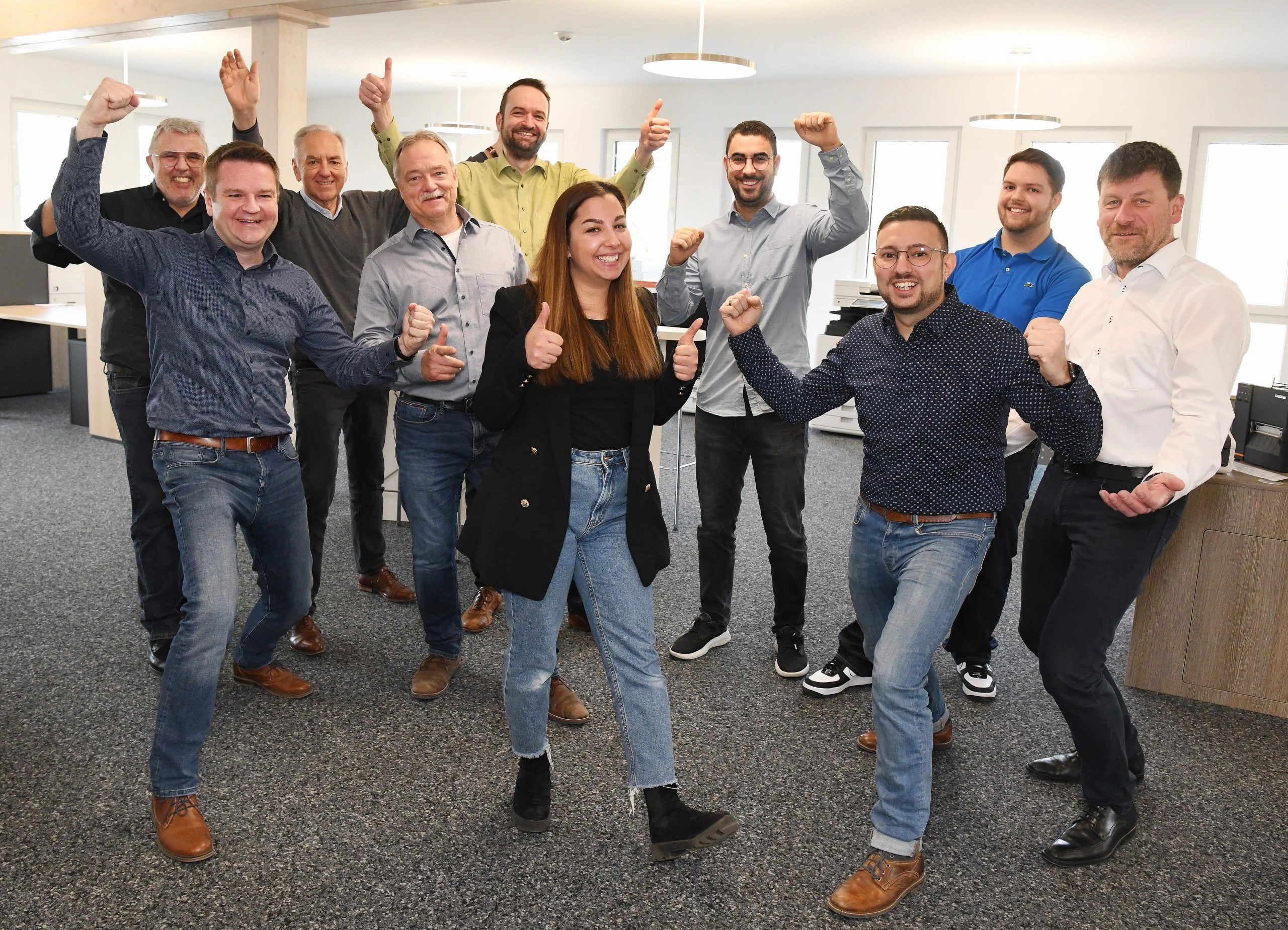 Eine Gruppe von fröhlichen Menschen steht in einem Büro und posiert ausgelassen, zeigt Daumen hoch.