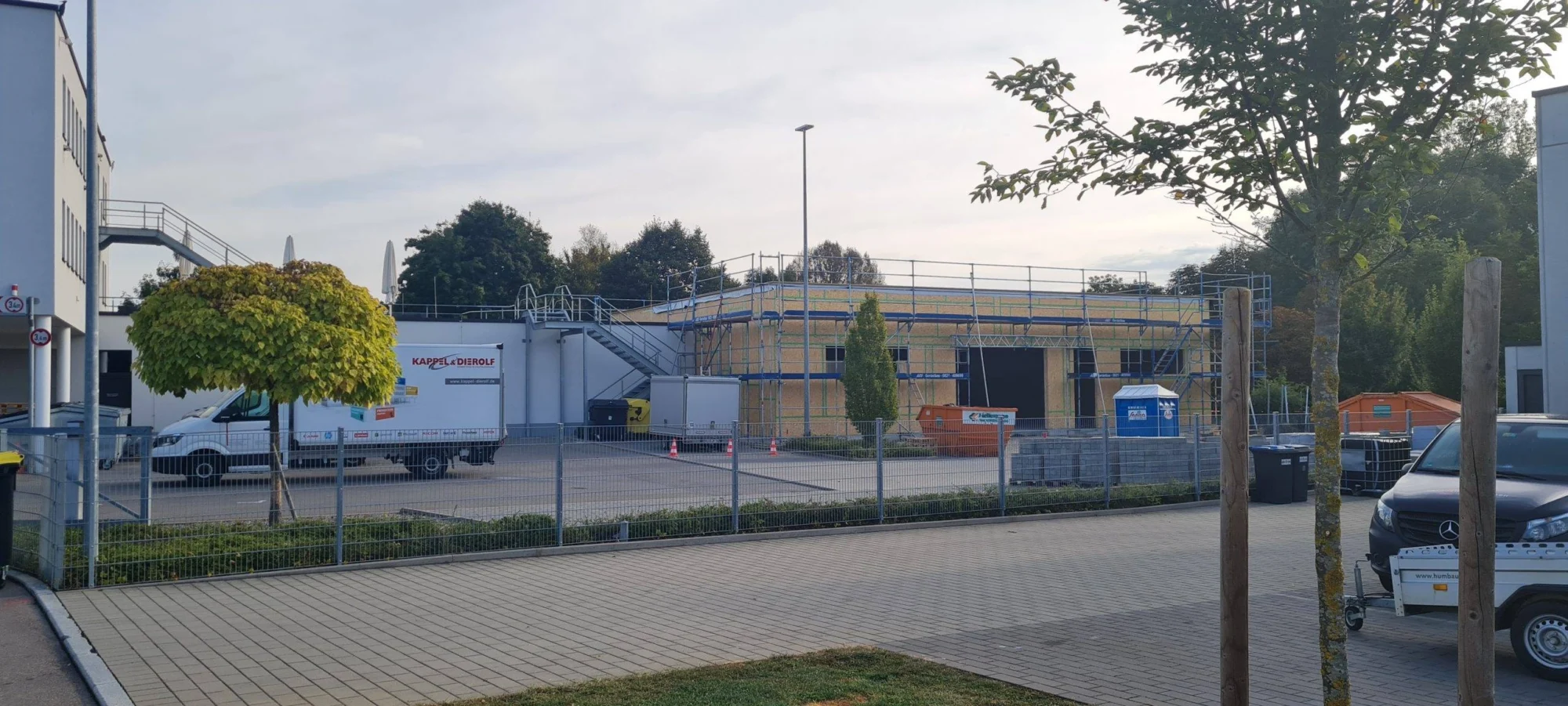 Bauplatz mit Gerüst und Lkw vor teilweise errichteter Halle, umgeben von Bäumen und Bauzäunen, bei hellem Tageslicht.