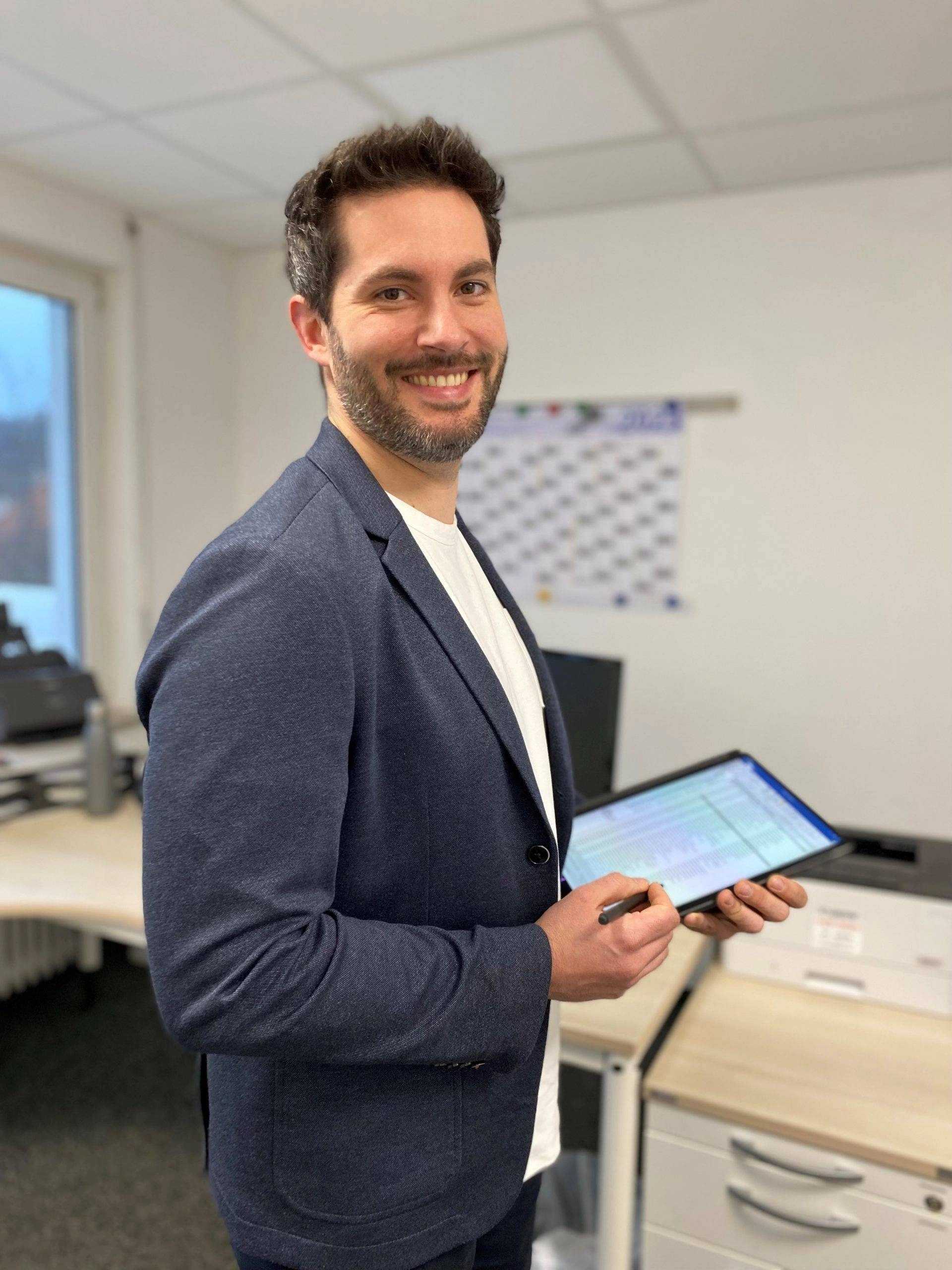 imgi_27_20230309_SS_Portrait-farbe-scaled Ein lächelnder Mann im Büro hält ein Tablet und Stift, steht vor einem Schreibtisch mit Drucker und Kalender an der Wand.