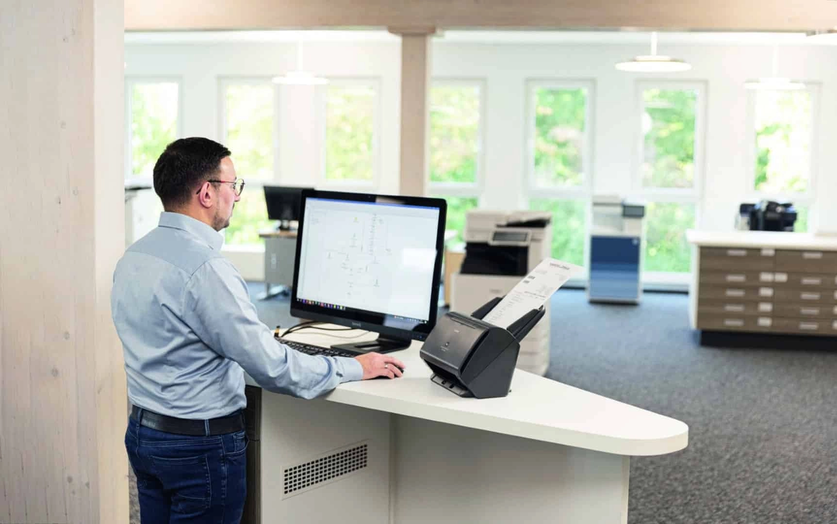 Mann arbeitet stehend am Computer mit Dokumentenscanner in modernem Büro, helle Umgebung mit Fenstern.