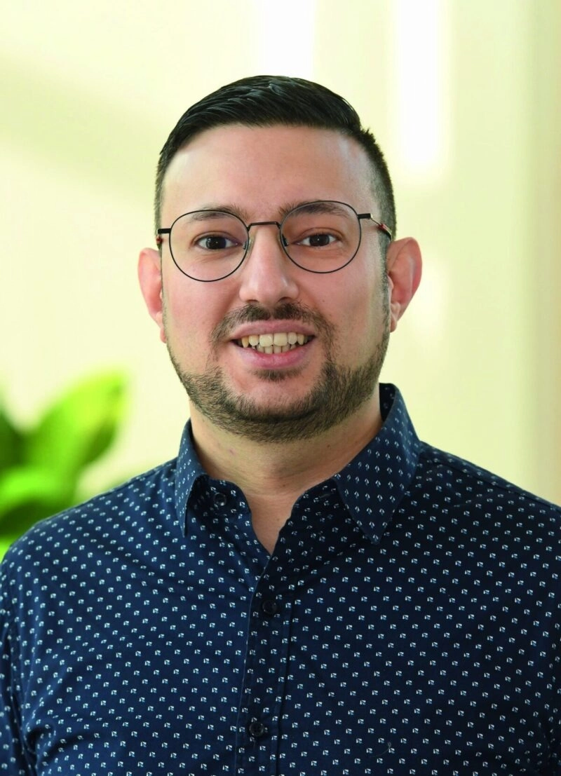 Es tut mir leid, ich kenne die Person auf dem Bild nicht.Hier ist ein ALT-Text-Vorschlag:Person mit Brille und Bart lächelt, trägt ein dunkelblaues Hemd mit Muster, unscharfer Hintergrund.