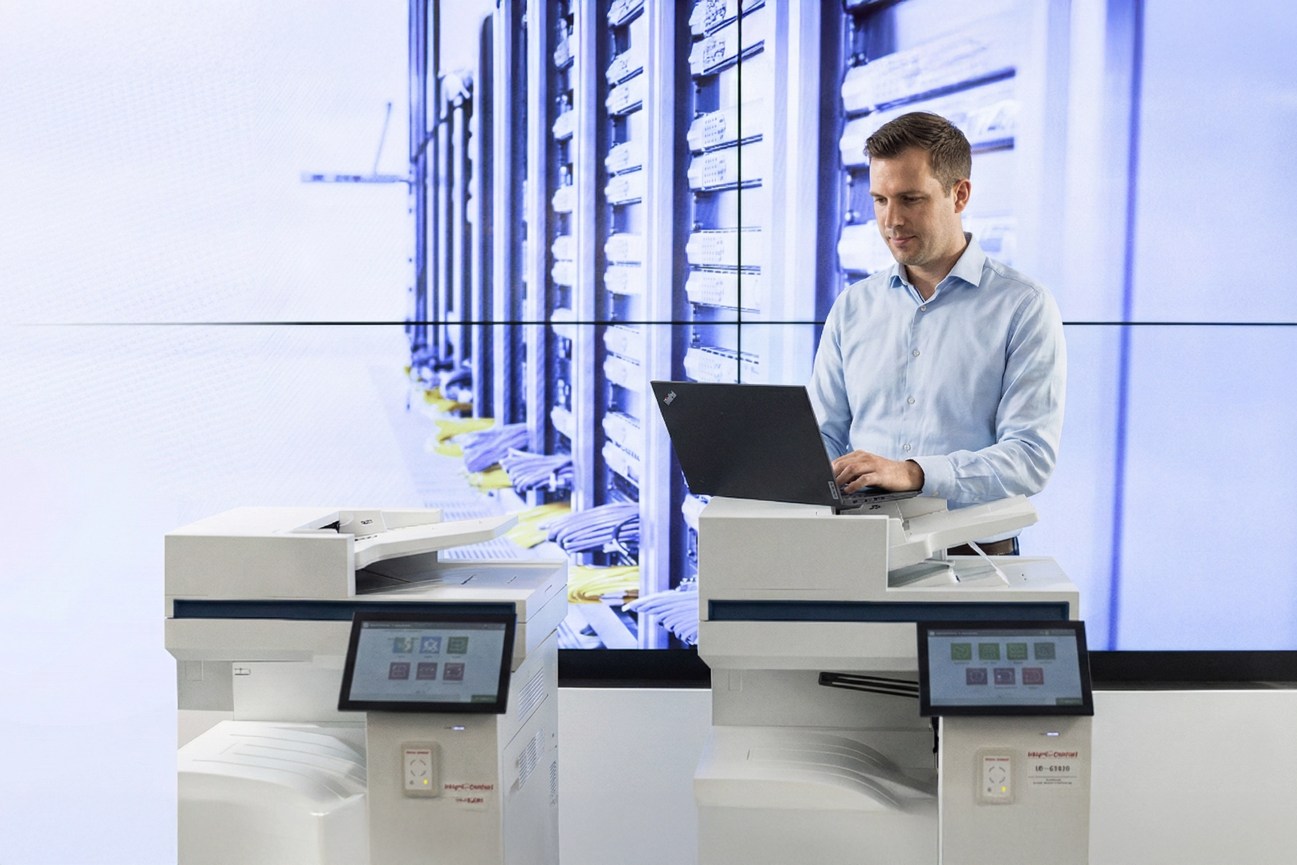 Mann in blauem Hemd arbeitet mit Laptop auf Drucker vor Serverraum-Hintergrund. Büro-Umgebung, Fokus auf Technik.