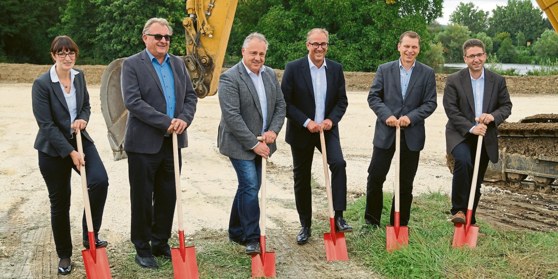 Sechs Personen in Anzügen halten Spaten bei feierlichem Spatenstich auf einer Baustelle im Freien.