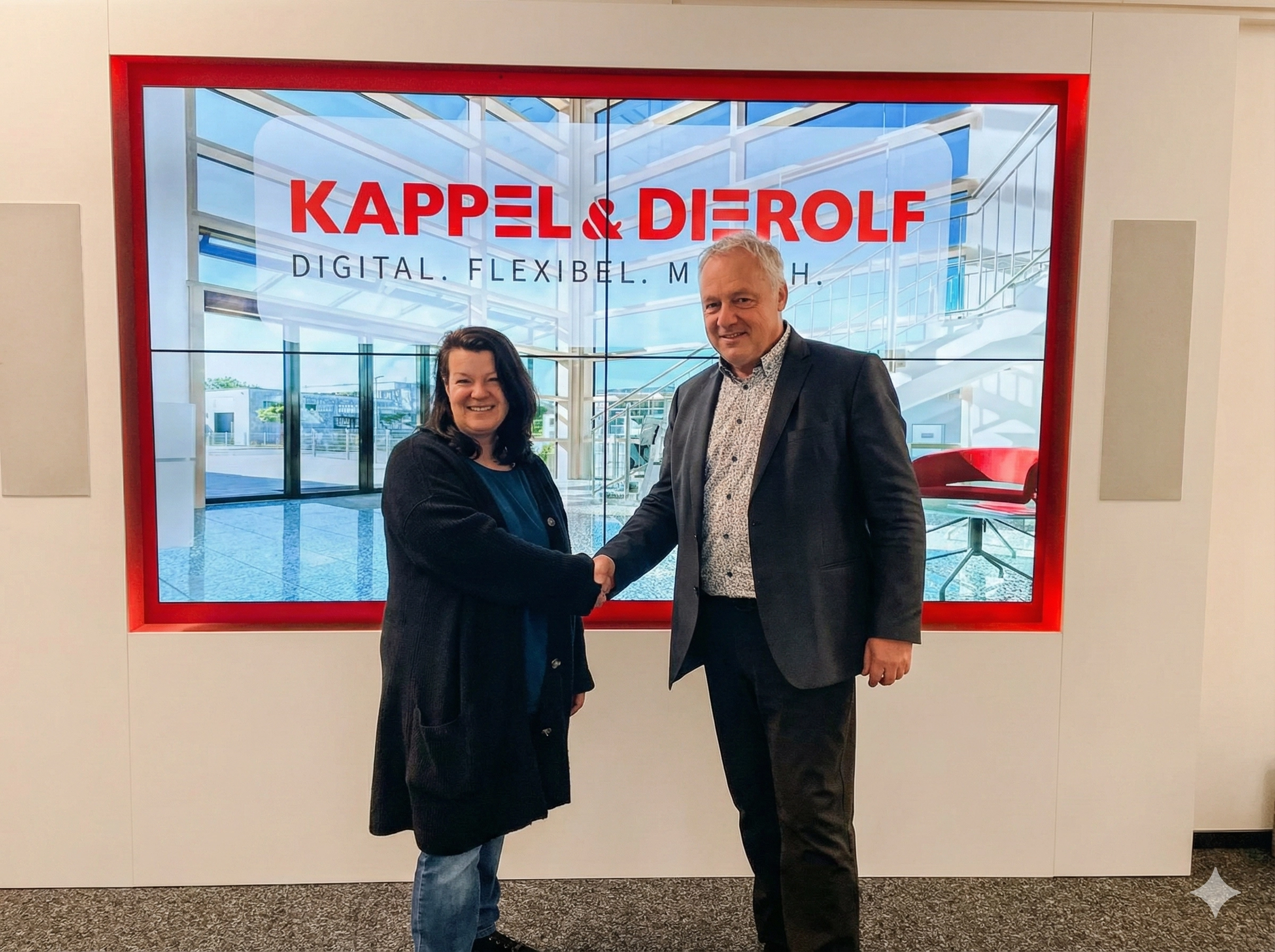 Zwei Personen schütteln Hände vor einem großen Bildschirm mit Kappel & Dierolf Werbung im Büro.