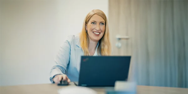 Eine Person in einem Büro sitzt lächelnd an einem Laptop, die Hand auf einer Computermaus ruhend.
