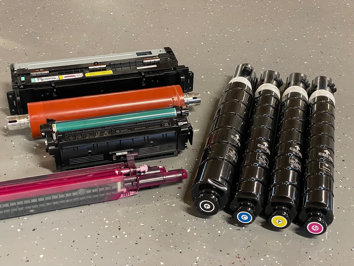 Verschiedene Druckerpatronen und Toner in Schwarz, Cyan, Gelb und Magenta auf einem grauen, gesprenkelten Boden.