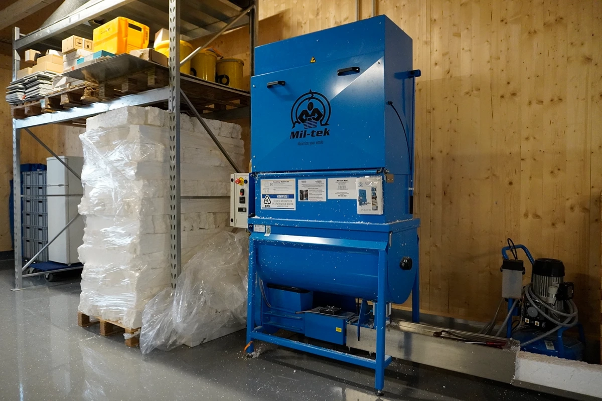 Blaue Müllpresse im Lager neben gestapeltem Verpackungsmaterial auf Paletten. Regale mit Kartons und Dosen darüber.
