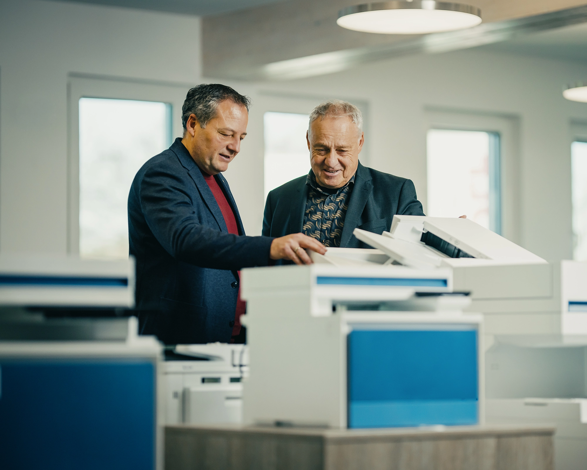 Zwei Männer betrachten zusammen einen Kopierer in einem Büro, diskutieren und zeigen auf das Gerät.