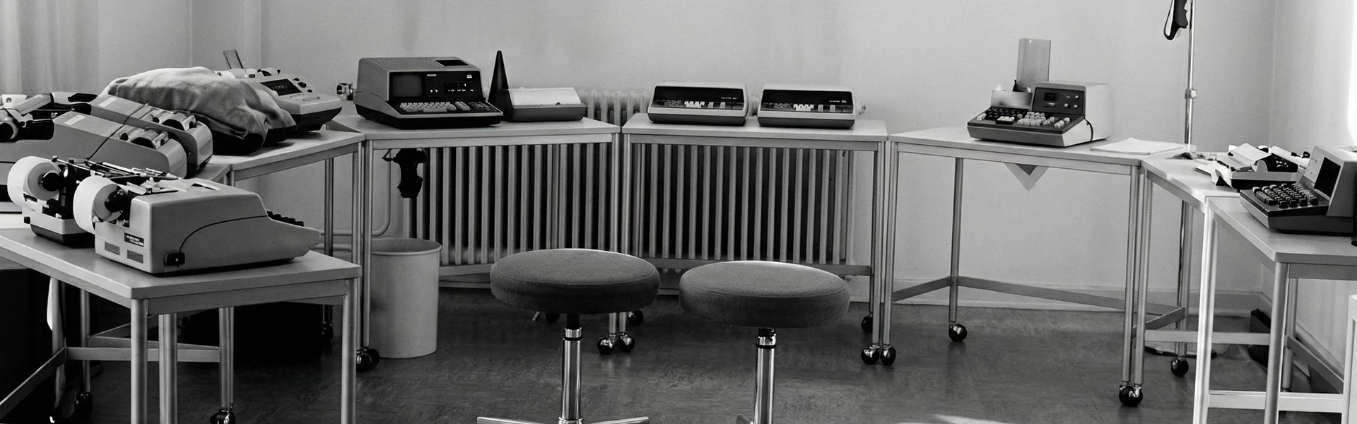 Schwarz-Weiß-Büro mit Schreibmaschinen und Rechenmaschinen auf Tischen, zwei gepolsterte Hocker im Vordergrund.