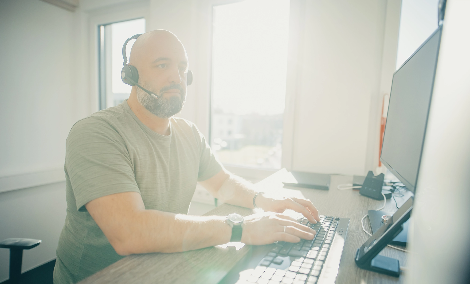 Mann mit Headset arbeitet an einem Computer in einem Büro, Licht strömt durch das Fenster.