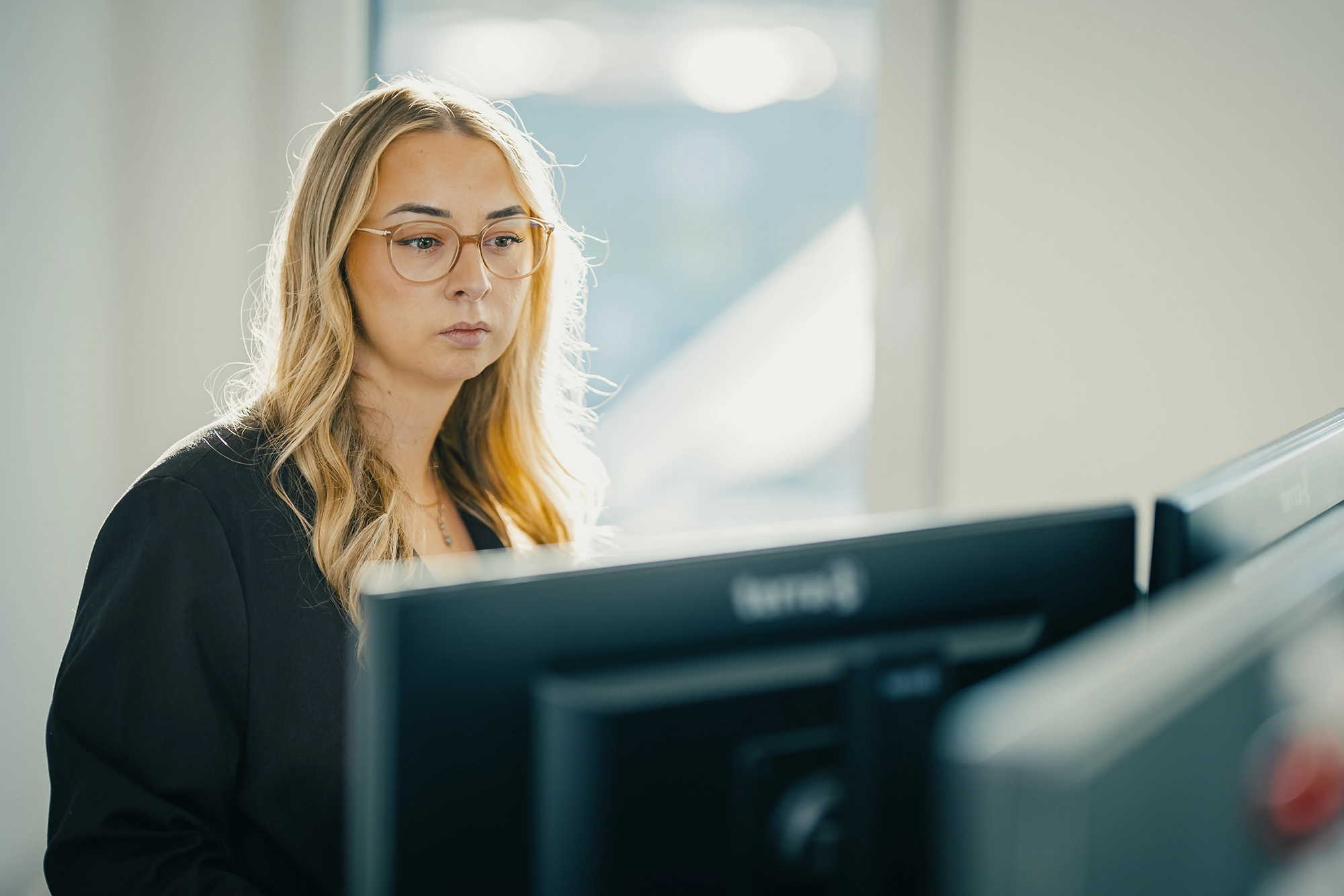 Frau mit Brille arbeitet konzentriert an zwei Monitoren in einem Büro mit natürlichem Licht.