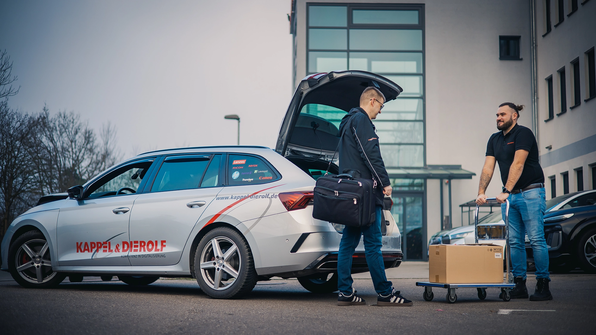 Zwei Männer verladen Pakete in ein Auto mit Firmenlogo, vor einem Bürogebäude. Einer nutzt einen Transportwagen.