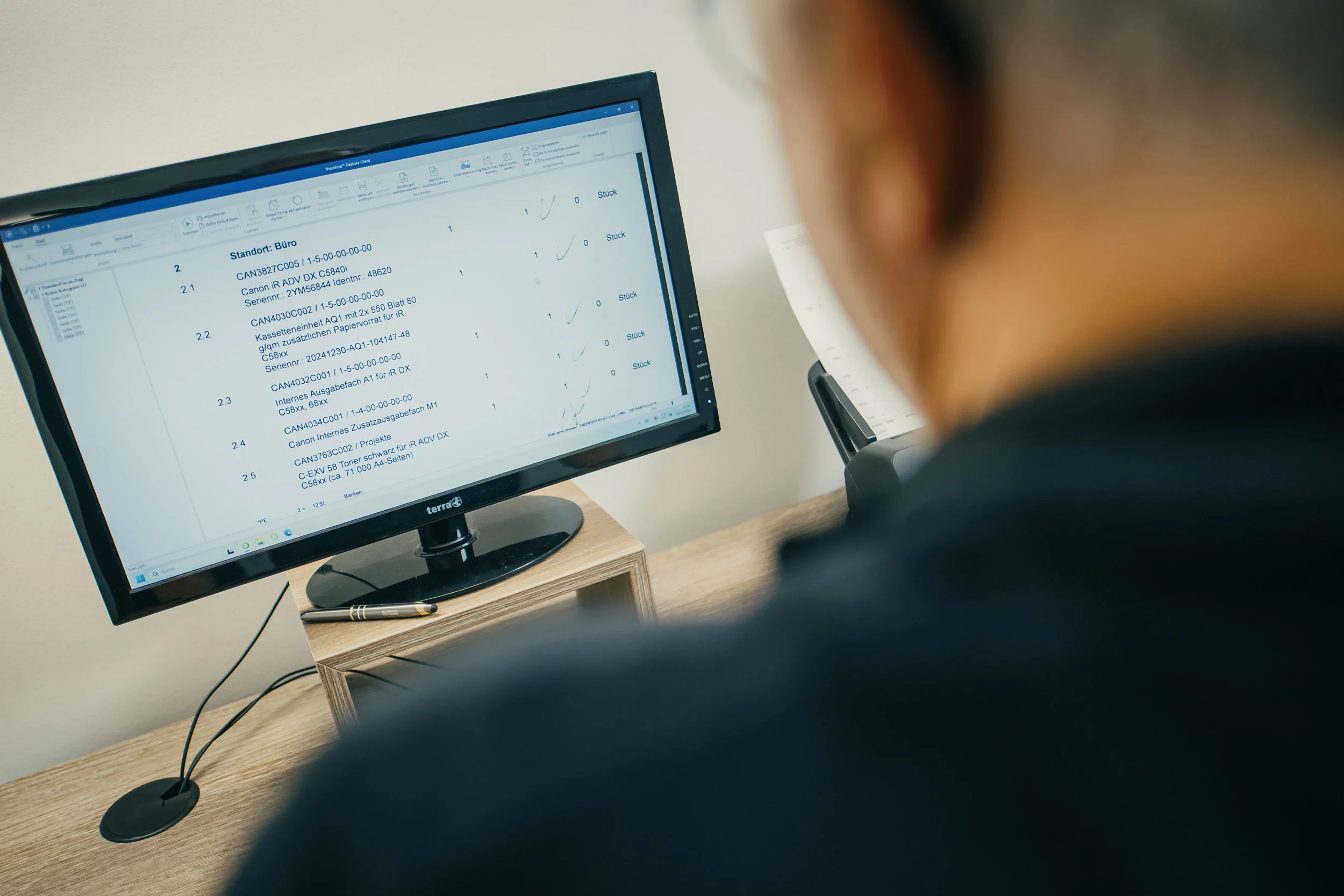 Eine Person betrachtet auf einem Monitor in einem Büro eine Liste technischer Informationen. Ein Stift liegt daneben.