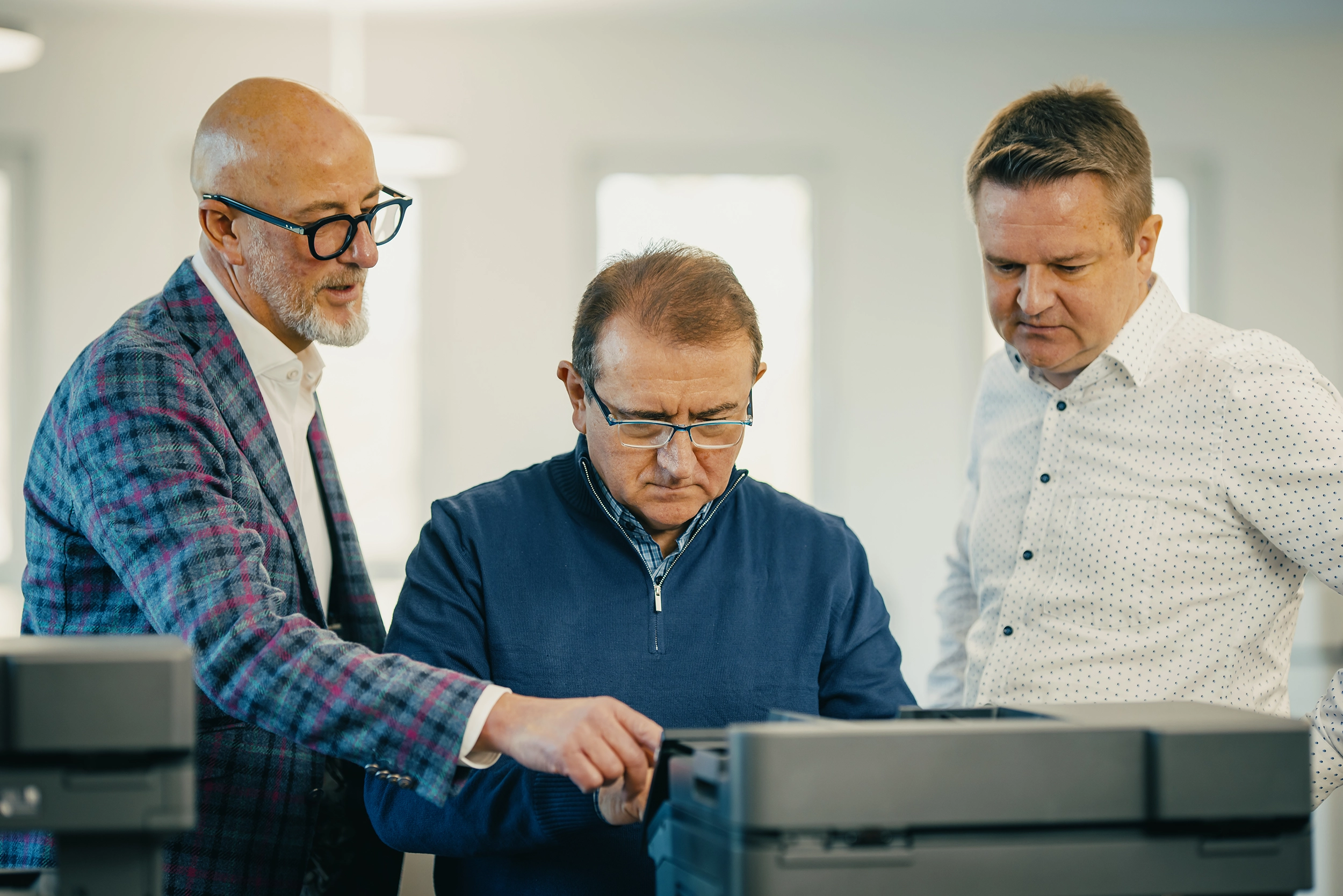 Drei Männer arbeiten konzentriert an einem Drucker in einem Büro, einer zeigt eine Anpassung.