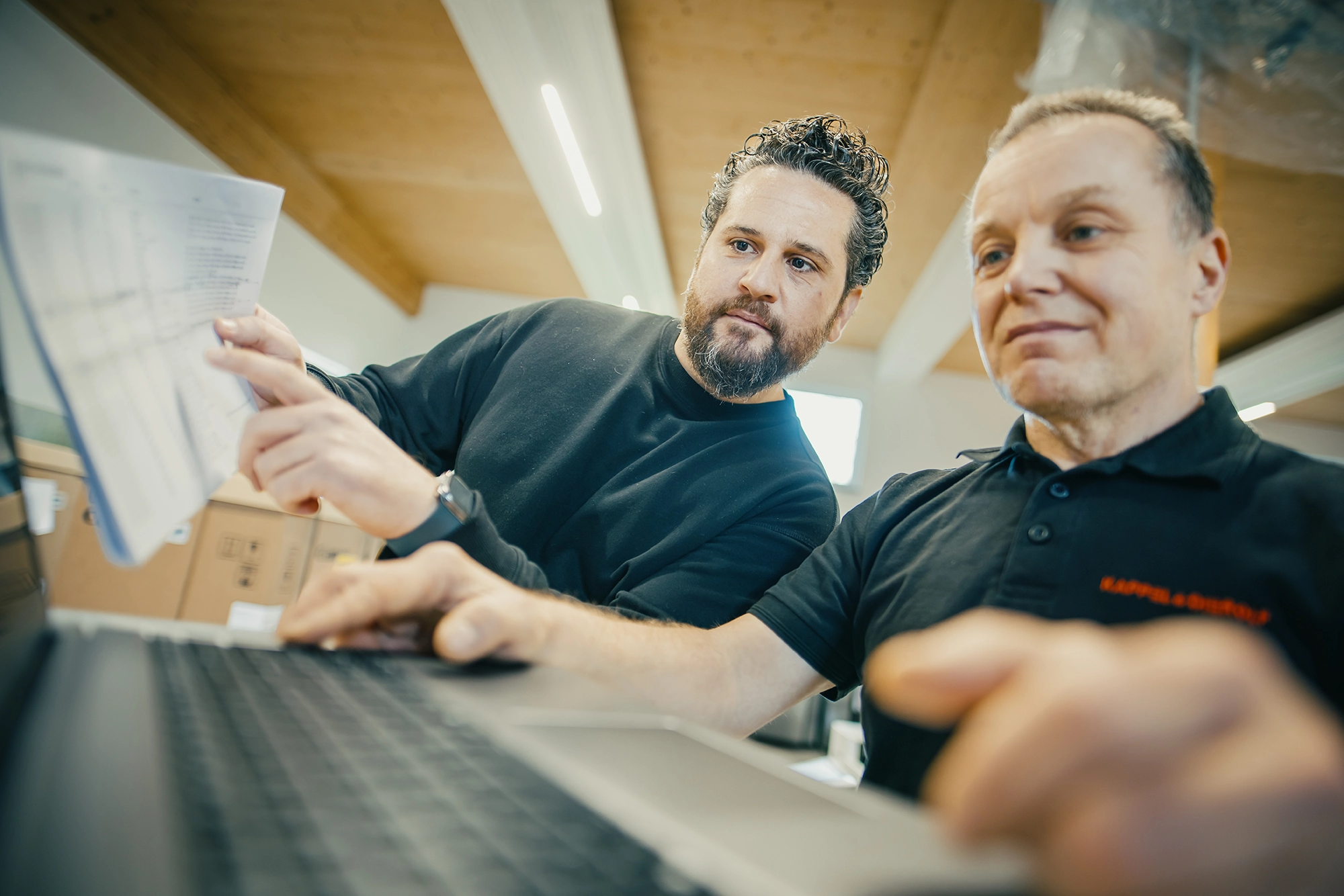 Zwei Männer arbeiten konzentriert an einem Laptop in einem Büro, einer hält Unterlagen.
