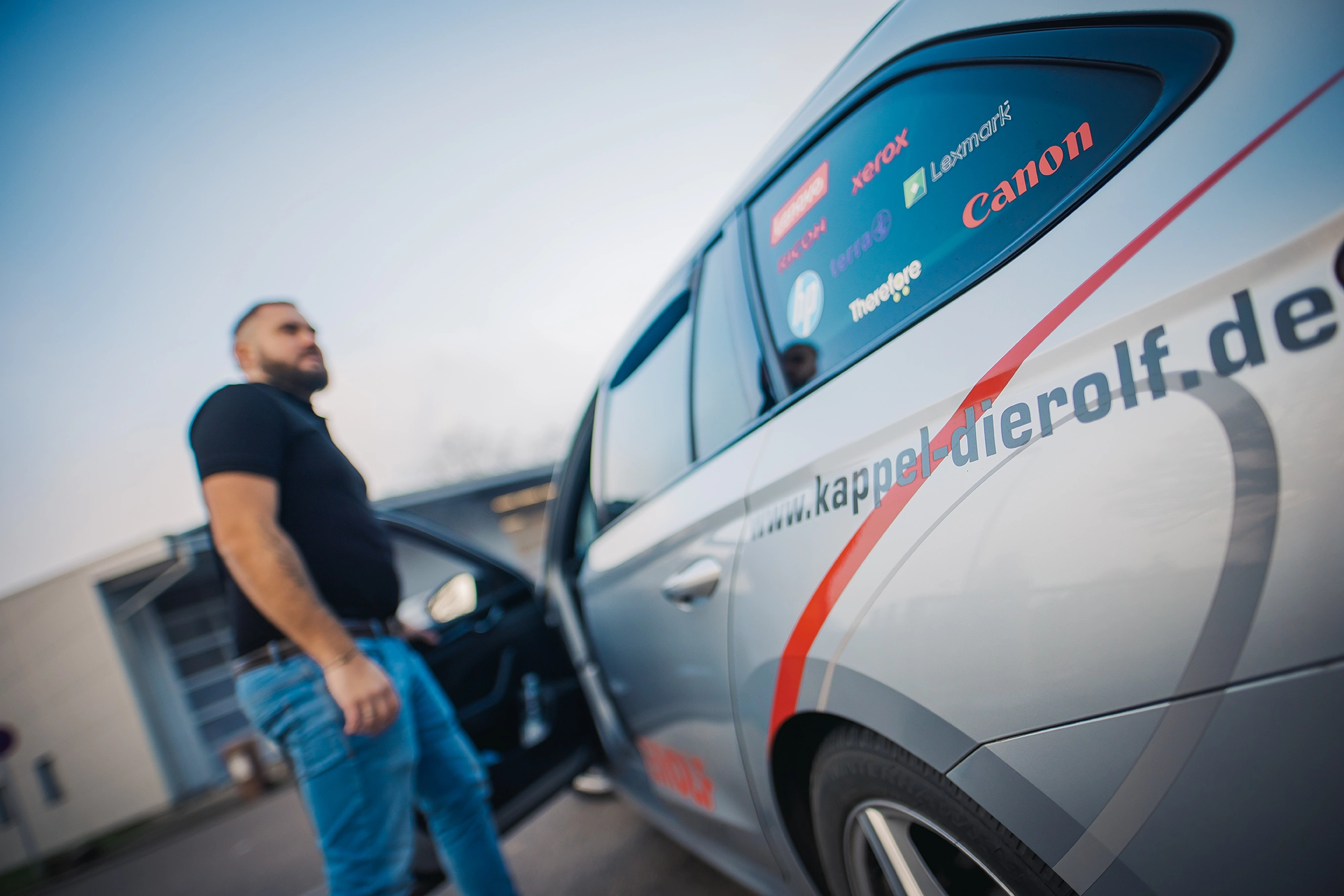 Kappel_Kompetenzen_Partner Ein Mann steht neben einem grauen Auto mit Werbung und geöffneter Tür; im Hintergrund ein Industriegebäude.