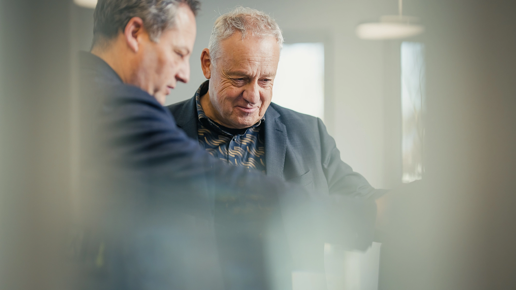 Zwei Männer im Anzug besprechen etwas in einem Büro, einer zeigt auf ein Dokument.
