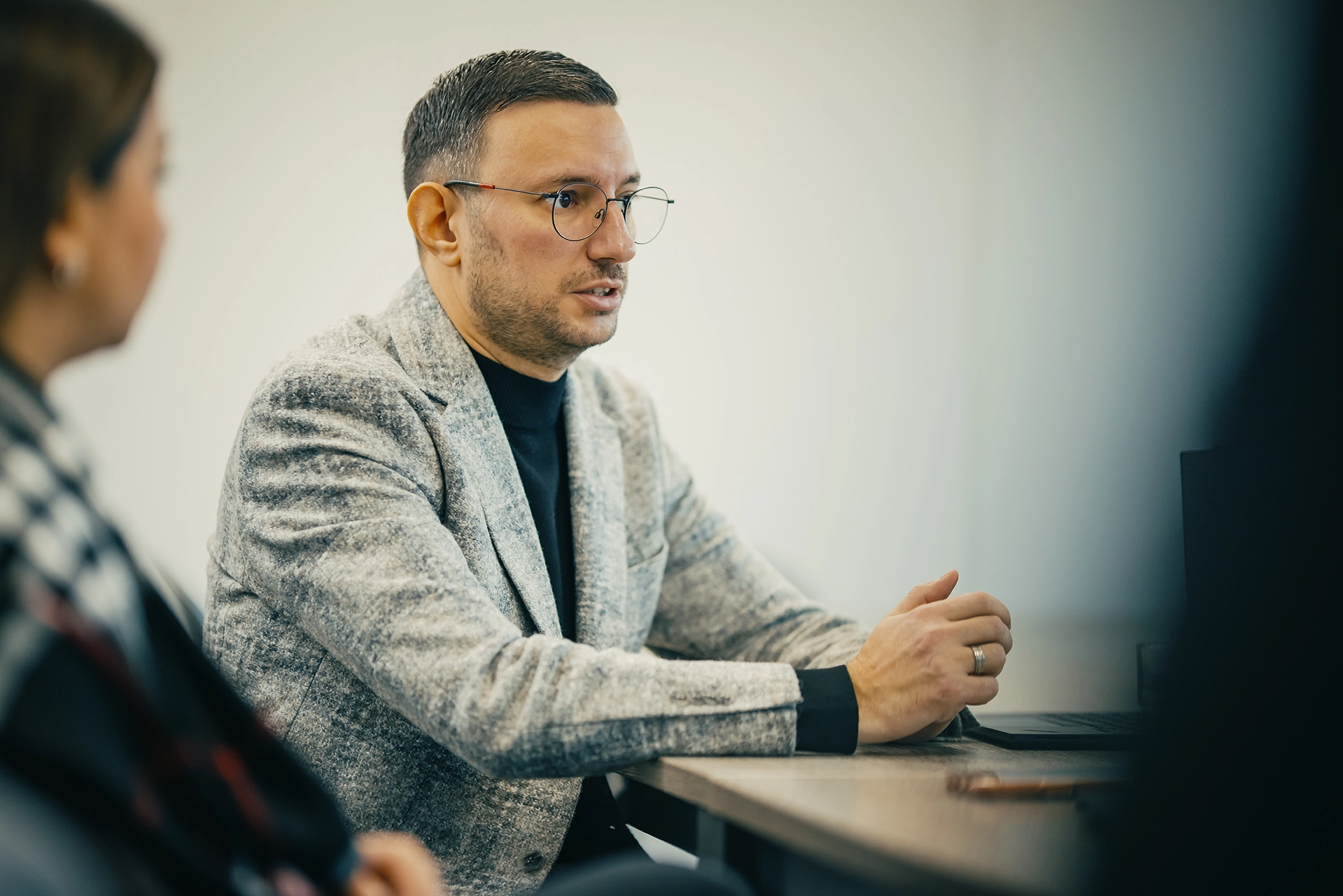 Mann und Frau im Gespräch an einem Tisch, Mann trägt Brille und grauen Blazer, Frau halb sichtbar.