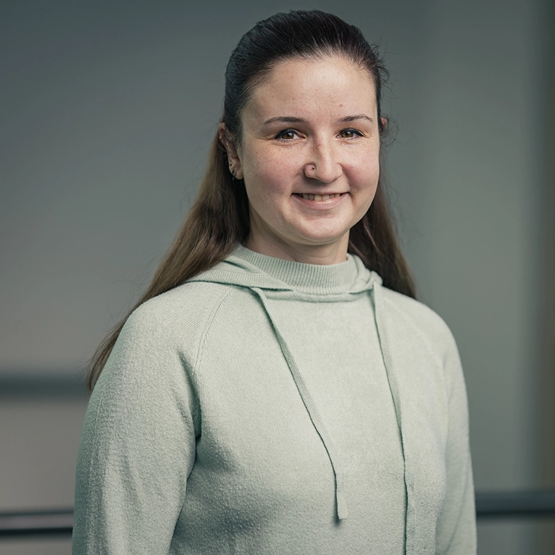Eine lächelnde Person in einem hellen Hoodie steht vor einem neutralen Hintergrund.
