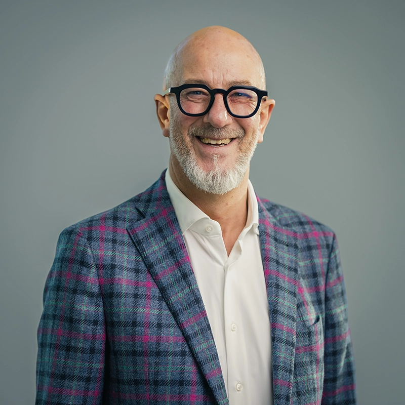 Kappel_Potrait_Döttinger Ein lächelnder Mann mit Brille, Glatze und Bart trägt ein kariertes Sakko vor neutralem Hintergrund.
