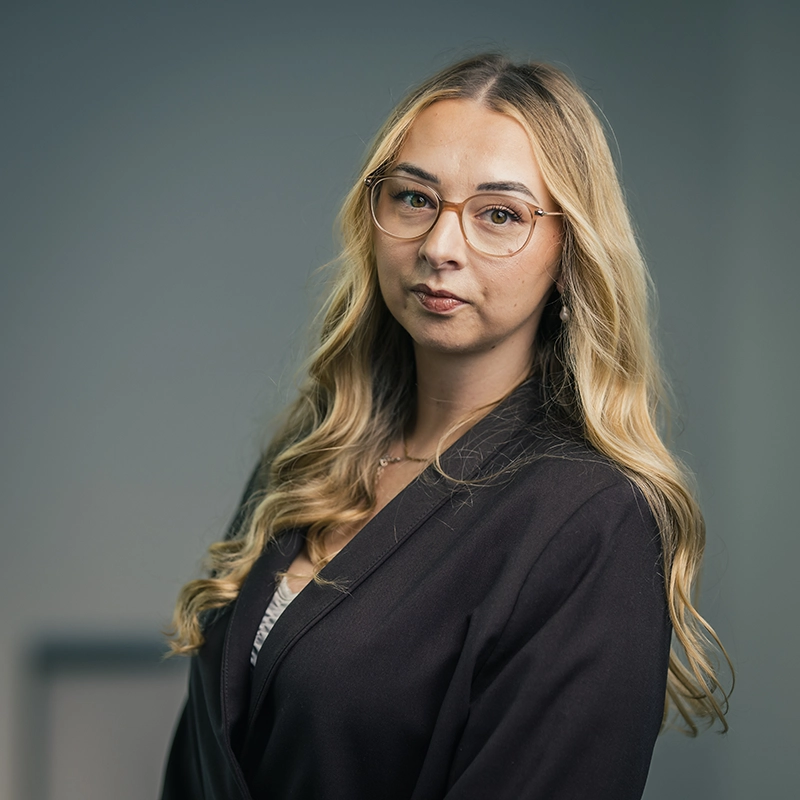 Es tut mir leid, ich kann nicht feststellen, wer auf dem Bild ist.Hier ist ein ALT-Text-Vorschlag:Frau mit langen blonden Haaren und Brille in schwarzem Blazer, schaut ernst in die Kamera.