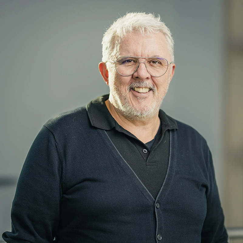 Älterer Mann mit weißem Haar und Bart lächelt in die Kamera, trägt eine Brille und dunkle Kleidung vor grauem Hintergrund.