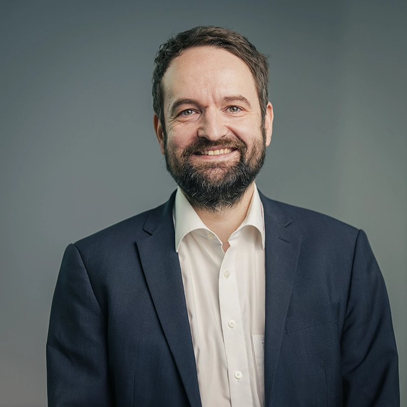 Ein lächelnder Mann in einem dunklen Anzug und weißem Hemd, vor grauem Hintergrund.