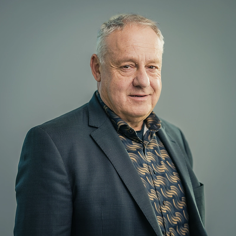 Kappel_Potrait_Fügel Eine Person im Anzug mit gemustertem Hemd schaut lächelnd in die Kamera, vor grauem Hintergrund.