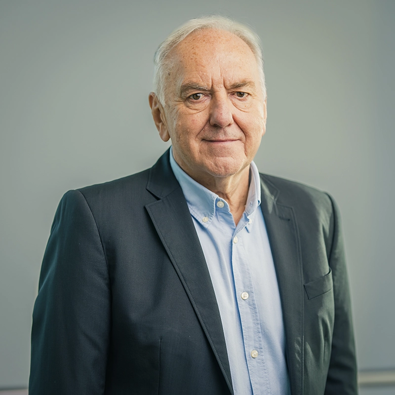 Älterer Mann in Anzug und Hemd posiert vor neutralem Hintergrund, schaut direkt in die Kamera.