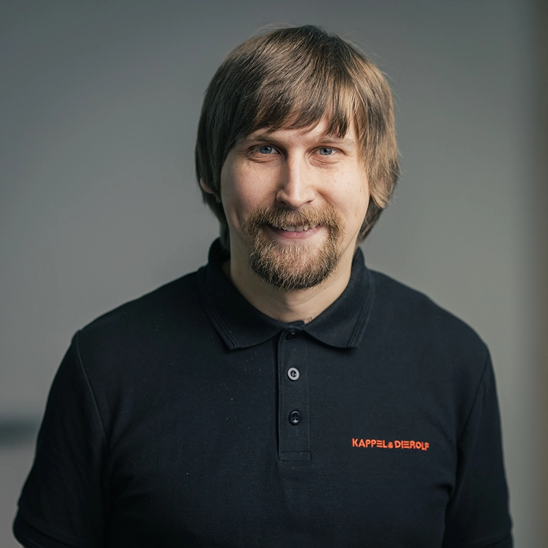 Es tut mir leid, ich kann nicht erkennen, wer auf dem Bild ist.Ein Mann in einem dunklen Polohemd mit Logo, lächelt vor neutralem Hintergrund.