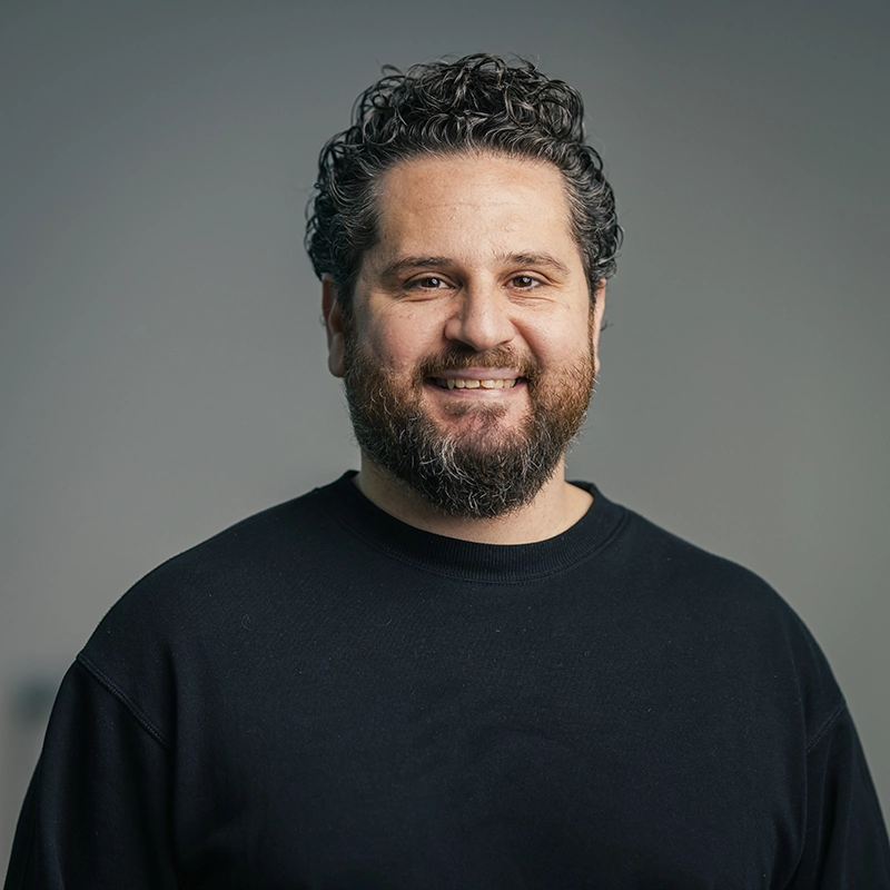 Es tut mir leid, ich kann diese Person nicht identifizieren.ALT-Text: Mann mit Bart und lockigem Haar lächelt vor grauem Hintergrund.