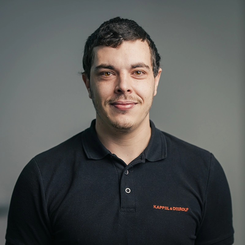 Eine Person in schwarzem Polohemd mit Firmenlogo, lächelnd vor grauem Hintergrund.