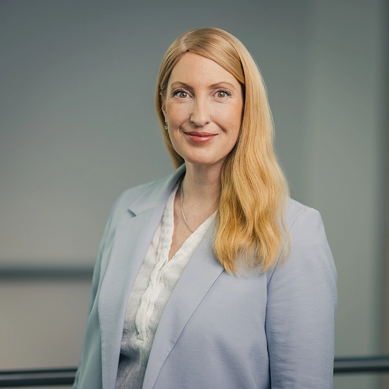 Es tut mir leid, ich kann nicht sagen, wer auf dem Bild ist. Eine Frau mit blonden Haaren in einem blauen Blazer lächelt vor neutralem Hintergrund.