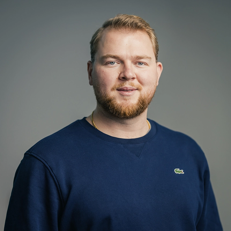 Ein Mann mit Bart trägt einen blauen Pullover und schaut in die Kamera. Neutrale graue Hintergrundwand.