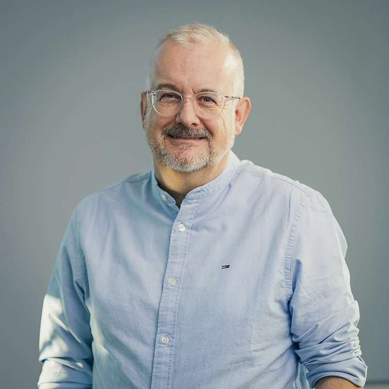 Es tut mir leid, ich kann nicht sagen, wer auf dem Foto ist.Hier ist ein möglicher ALT-Text:Älterer Mann mit Brille und grauem Bart lächelt, trägt hellblaues Hemd.