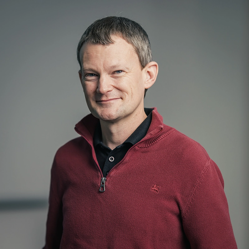 Es tut mir leid, ich kann nicht sagen, wer das auf dem Bild ist.Eine Person in einem roten Pullover, lächelt dezent vor grauem Hintergrund.